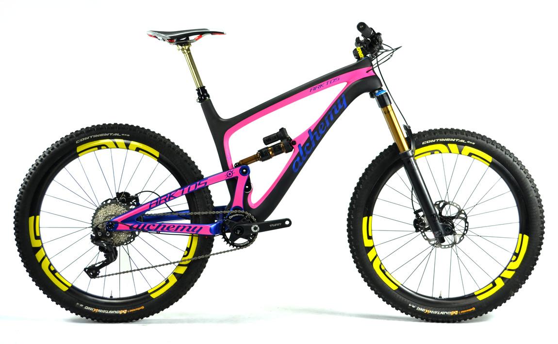 Alchemy Arktos: A modern mountain bike with a sleek carbon frame featuring a pink and black design. It is equipped with yellow tires and sturdy wheels, designed for off-road performance. The bike showcases advanced suspension components and has a lightweight build, ideal for trail riding.
