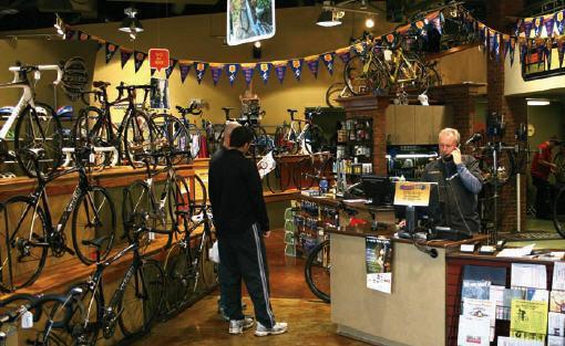 A bicycle shop interior featuring several bikes on display along the walls, with a customer conversing with a cashier at the front counter. Decorative banners are hung from the ceiling, and various cycling accessories are visible on the shelves.