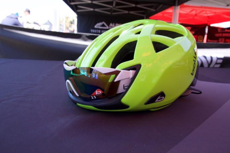 A bright green cycling helmet with aerodynamic design resting on a dark table, accompanied by a pair of sunglasses featuring mirrored lenses. The background includes blurred tent structures and event setups, suggesting an outdoor cycling event.
