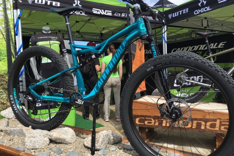 A brightly colored Cannondale mountain bike displayed at an outdoor event, with green tents and branding in the background. The bike features large tires and a detailed suspension system, and is positioned on a bed of rocks, showcasing its rugged design. People in cycling attire are partially visible in the background.