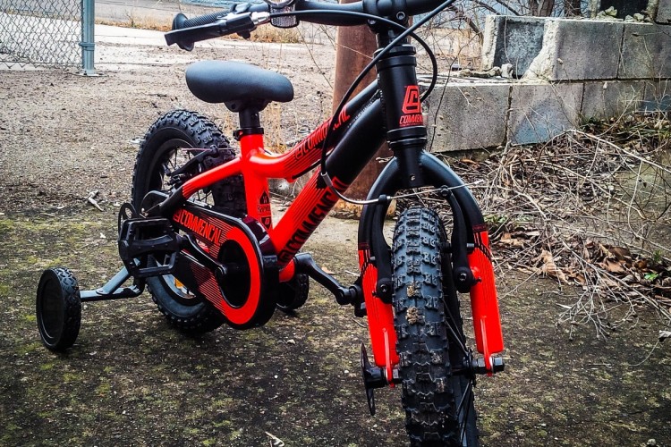 A small, bright red children's bicycle with training wheels is positioned on a gravel surface. The bike features a sturdy frame, knobby tires for off-road riding, and a small headlight mounted on the handlebars. In the background, a chain-link fence and a gray building can be seen, along with some scattered leaves and an overcast sky.