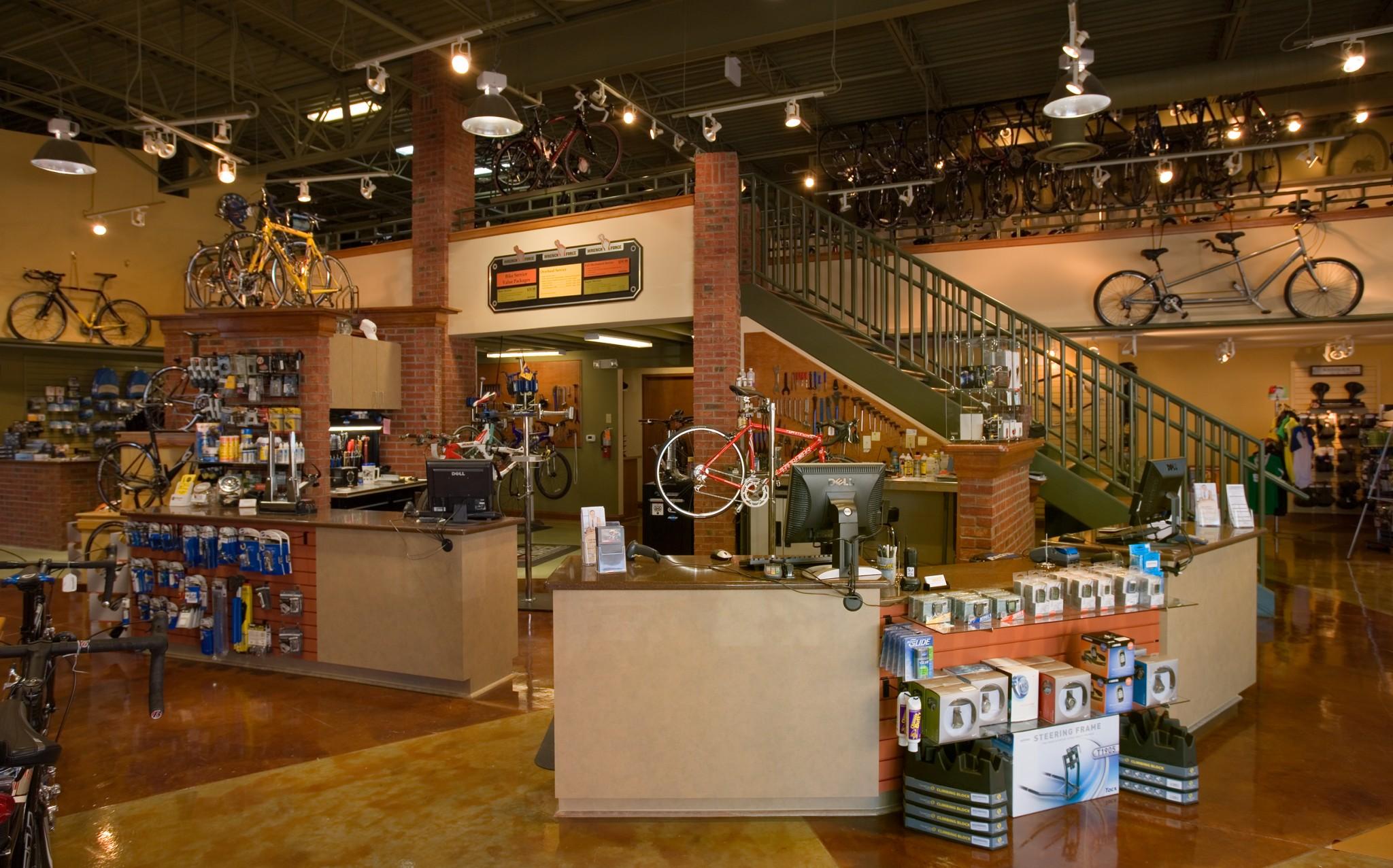 Interior of a bicycle shop featuring a contemporary layout. The space includes a customer service area with multiple checkout counters, bikes displayed on walls and ceilings, and various bicycle accessories and tools on shelves. A staircase leads to a second level with additional bike displays, creating a vibrant, inviting atmosphere for cycling enthusiasts.