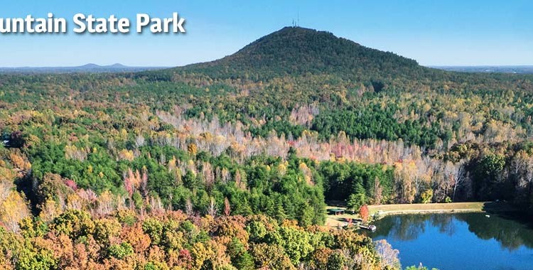 Aerial view of Crowders Mountain State Park, showcasing a lush landscape with vibrant fall foliage, a serene lake at the base, and the prominent Crowders Mountain in the background under a clear blue sky.