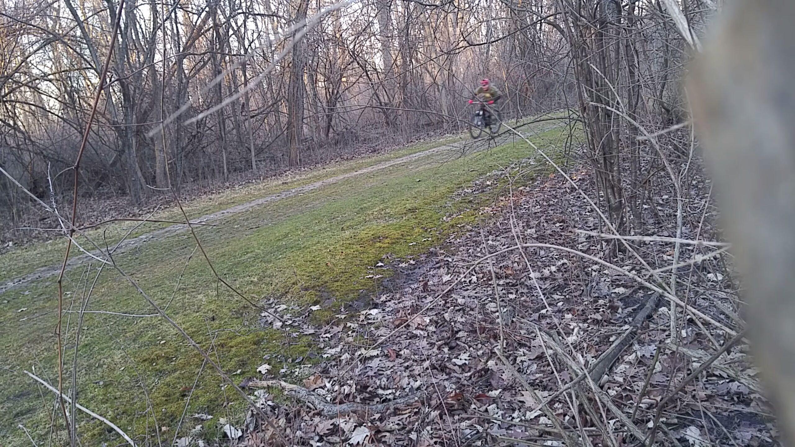 A cyclist riding along a narrow trail surrounded by bare trees and autumn leaves, with patches of grass visible on the ground. The scene captures a serene moment in a natural setting, suggesting the transition from winter to spring. Elizabeth Park Clydesdale Loop mountain bike trail.