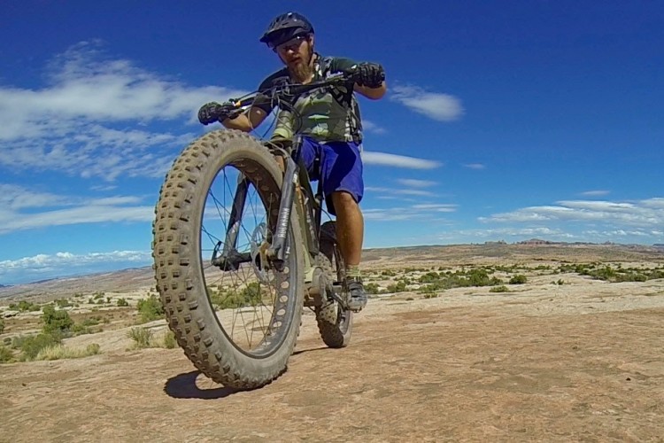 A mountain biker performing a stunt on rocky terrain, with the front wheel lifted off the ground. The rider is wearing a helmet and cycling gear, and the landscape features blue skies with clouds and distant mountains.