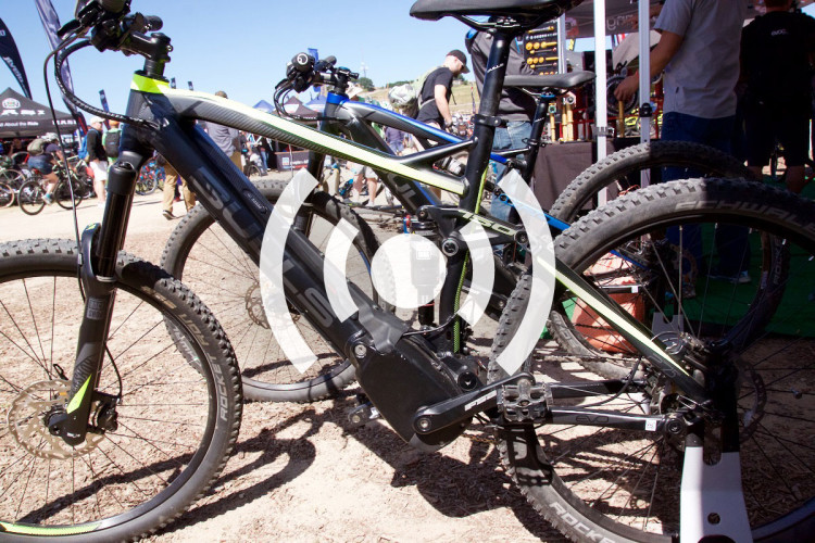 An electric mountain bike is showcased at an outdoor event, with a focus on the bike's mid-drive motor and components. In the background, various people are interacting and looking at other bikes. The setting is bright and sunny, highlighting the event's energetic atmosphere.