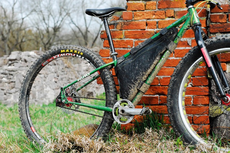 A green mountain bike with a black frame bag is leaning against a weathered brick wall. The bike features rugged tires, and there are patches of grass and dirt around it. The background shows remnants of a stone structure, indicating an outdoor, possibly rustic setting.