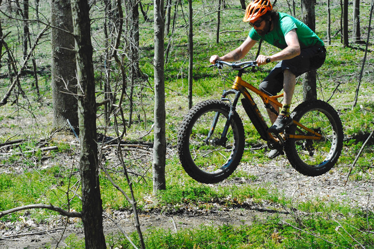 A person riding a fat bike in a wooded area, captured mid-jump with both wheels off the ground. The rider is wearing an orange helmet and a green shirt, with visible dirt on their legs, indicating an adventurous ride through the forest. Surrounding trees and green foliage provide a natural backdrop.