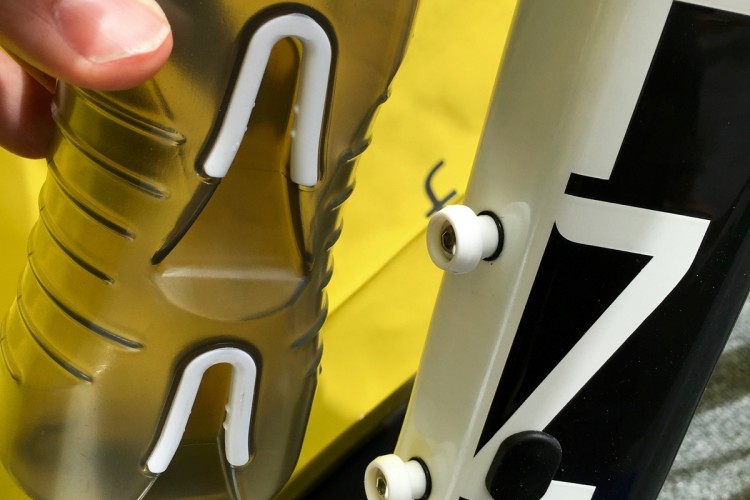 A close-up image of a hand holding a clear sports water bottle with a white grip, positioned next to a bicycle frame with visible mounting attachments. The background features a yellow surface, highlighting the scene.