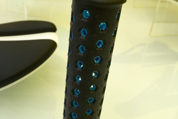 A close-up image of a black rubber grip handle with hexagonal cutouts and blue accents. The handle stands upright on a smooth surface, surrounded by bicycle or sports equipment in the background. The background features a bright yellow color, enhancing the focus on the grip.