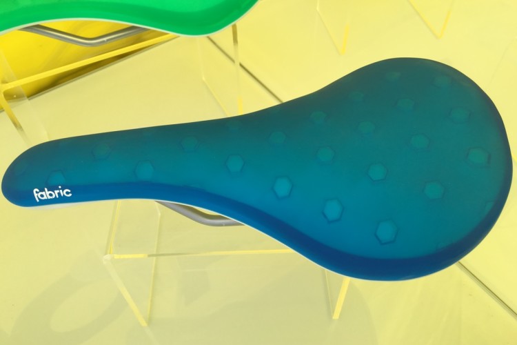 Blue bicycle saddle with a textured hexagonal pattern, displayed on a clear stand against a yellow background. The brand name "fabric" is visible on the side.