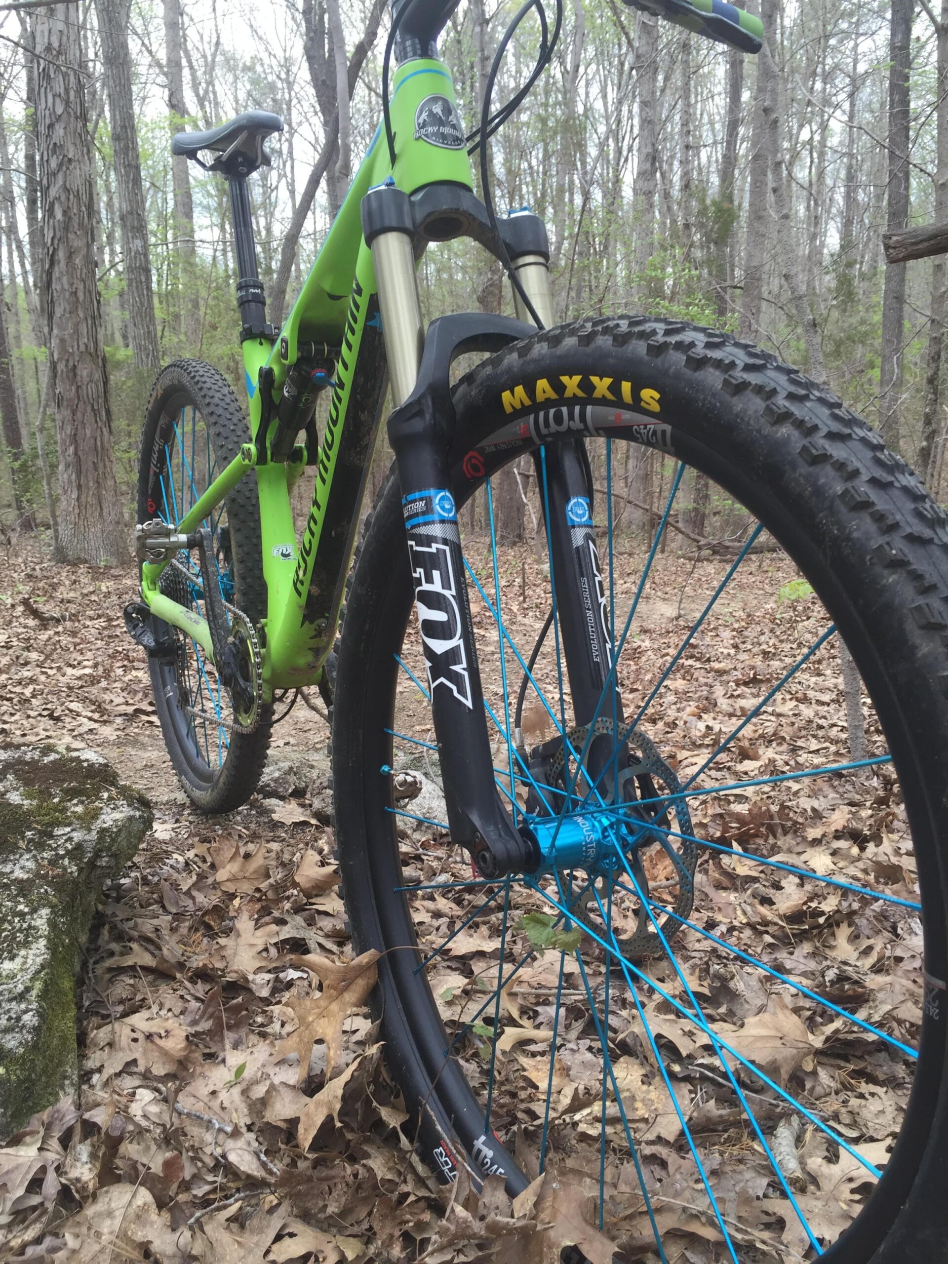 Rocky Mountain Instinct 950 MSL: A close-up view of a bright green mountain bike positioned on a forest path covered in brown leaves. The bike features a black front fork with blue accents and a prominent Maxxis tire. Trees in the background suggest an outdoor setting, ideal for mountain biking.