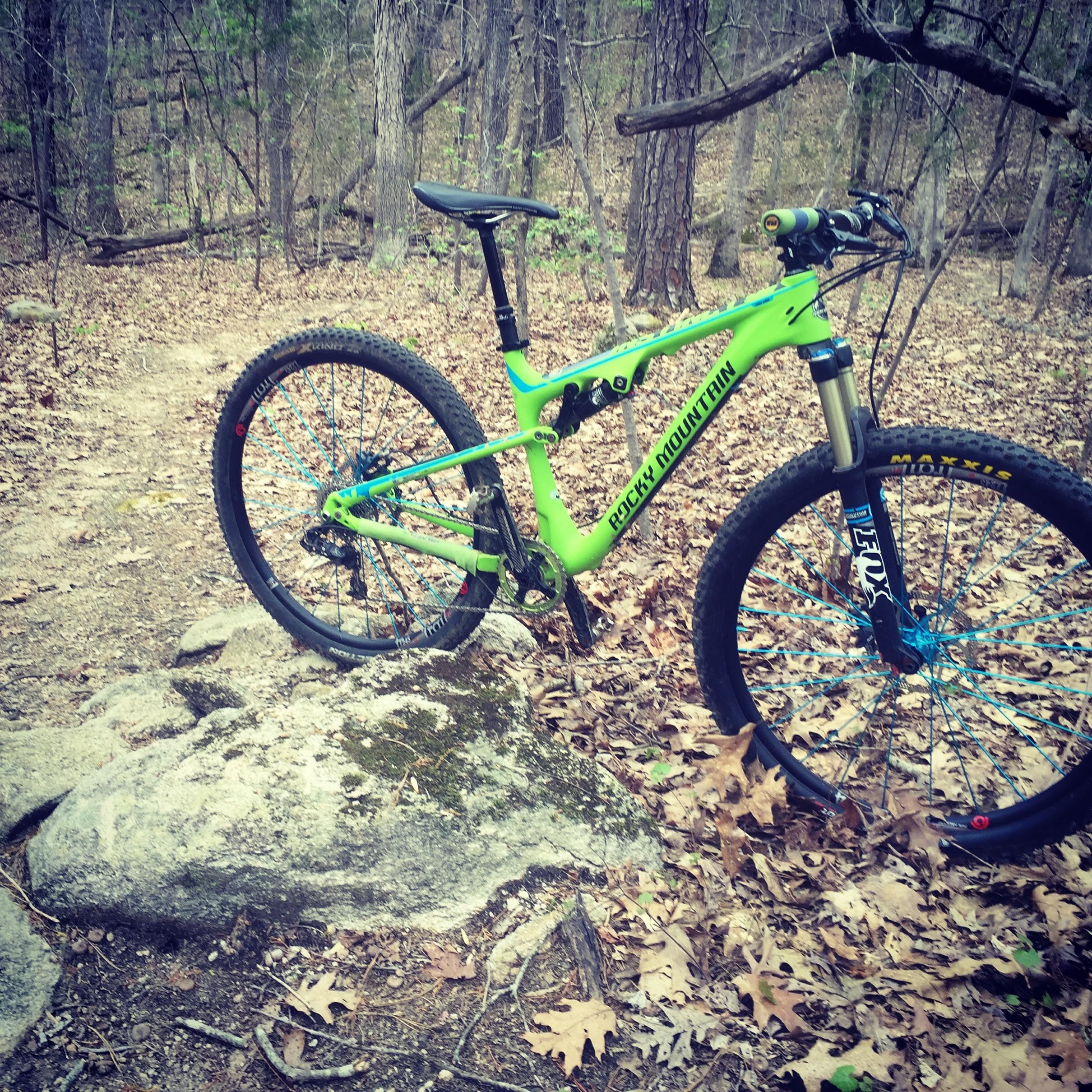 Rocky Mountain Instinct 950 MSL: A bright green mountain bike is positioned on a rocky trail surrounded by trees and fallen leaves in a wooded area. The bike features a sleek design with blue accents and fat tires, indicating it is ready for off-road cycling adventures.