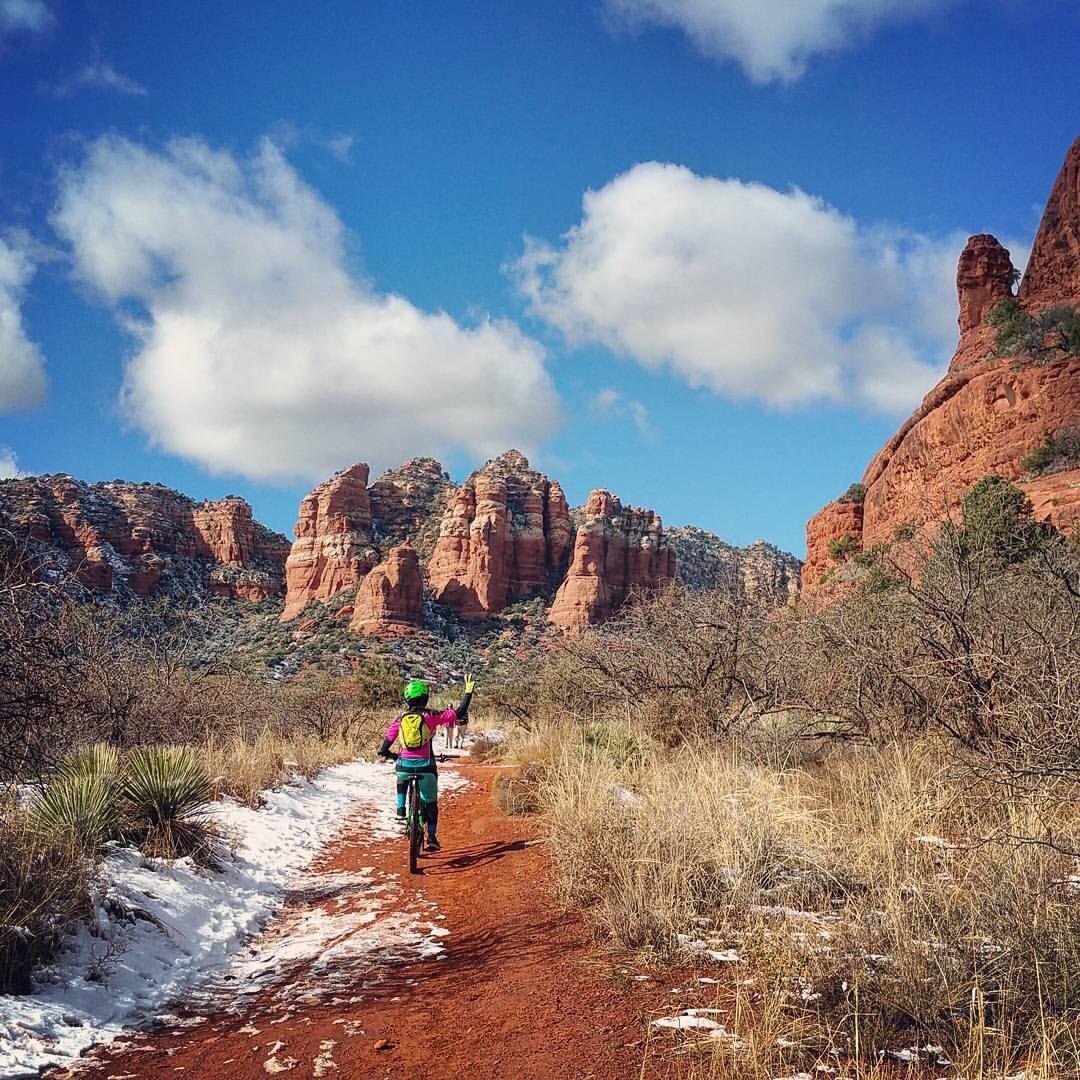 A cyclist wearing a bright green helmet and colorful jacket rides along a dirt path surrounded by red rock formations and sparse vegetation. Snow is visible on the ground, and there are fluffy white clouds in the blue sky. The scene captures a moment of adventure in a scenic outdoor setting. Bell Rock Trailway mountain bike trail.