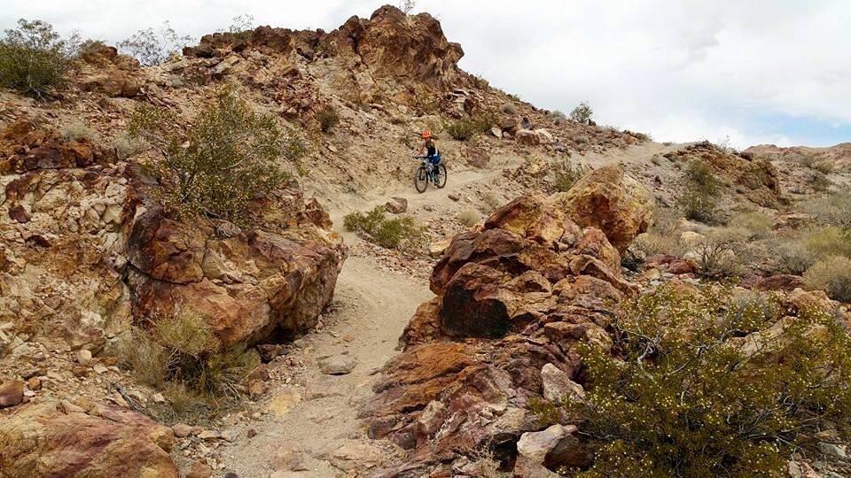 A cyclist navigating a rocky mountain trail surrounded by rugged terrain and sparse vegetation under a cloudy sky. Bootleg Canyon mountain bike trail.