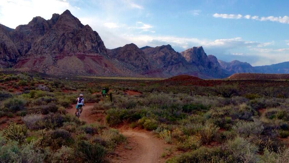 Two mountain bikers riding along a dirt trail in a desert landscape, surrounded by rocky mountains and sparse vegetation under a partly cloudy sky. Blue Diamond mountain bike trail.