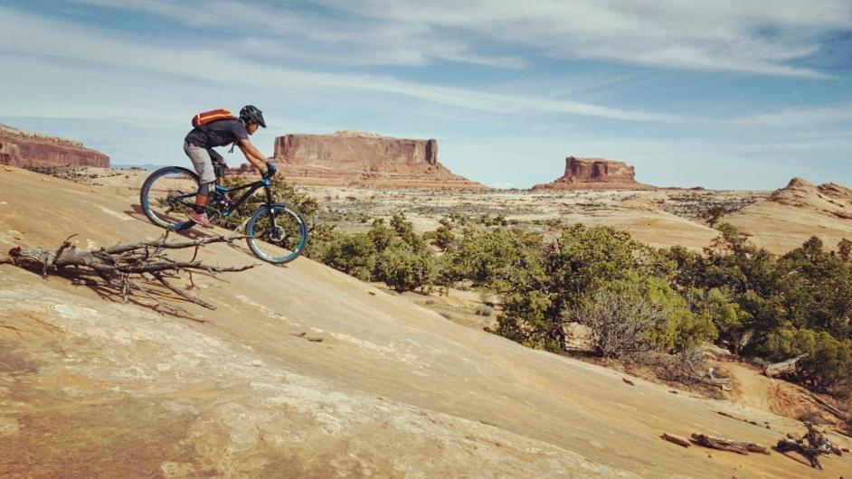 A mountain biker skillfully rides down a rocky slope, navigating over a fallen branch. In the background, majestic rock formations and a wide landscape are visible under a blue sky with scattered clouds. Navajo Rocks mountain bike trail.