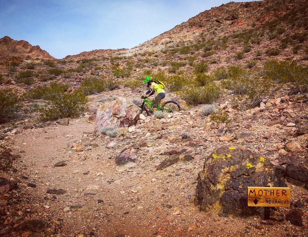 A mountain biker navigates a rocky trail in a desert landscape, with a sign indicating "Mother" trail, marked as "Advanced." The cyclist is wearing a bright yellow shirt and a green helmet, surrounded by sparse vegetation and rugged terrain under a blue sky. Bootleg Canyon mountain bike trail.