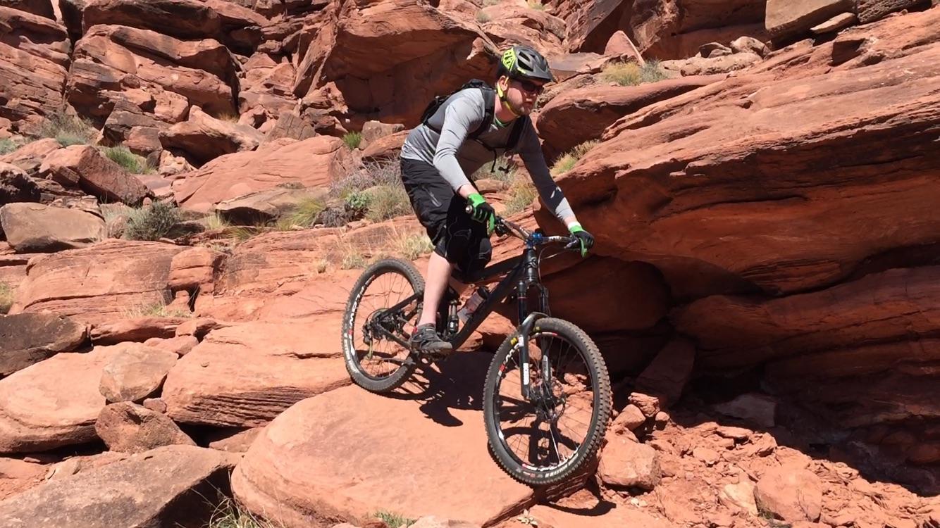 A mountain biker navigating rocky terrain, surrounded by red rock formations and sparse vegetation. The rider is wearing a helmet and protective gear, focused on maintaining balance while riding over uneven surfaces. Captain Ahab mountain bike trail.