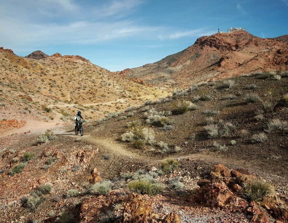 A mountain biker riding along a winding dirt trail in a rugged desert landscape. The scene features arid hills with varying shades of brown and red, scattered shrubs, and a clear blue sky above. Bootleg Canyon mountain bike trail.