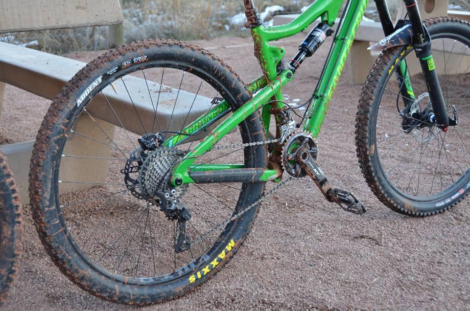 Santa Cruz Bronson: A close-up view of the rear section of a green mountain bike, showcasing its muddy tires and drivetrain components. The bike is resting on a sandy surface next to a wooden bench, indicating recent use in outdoor terrain.