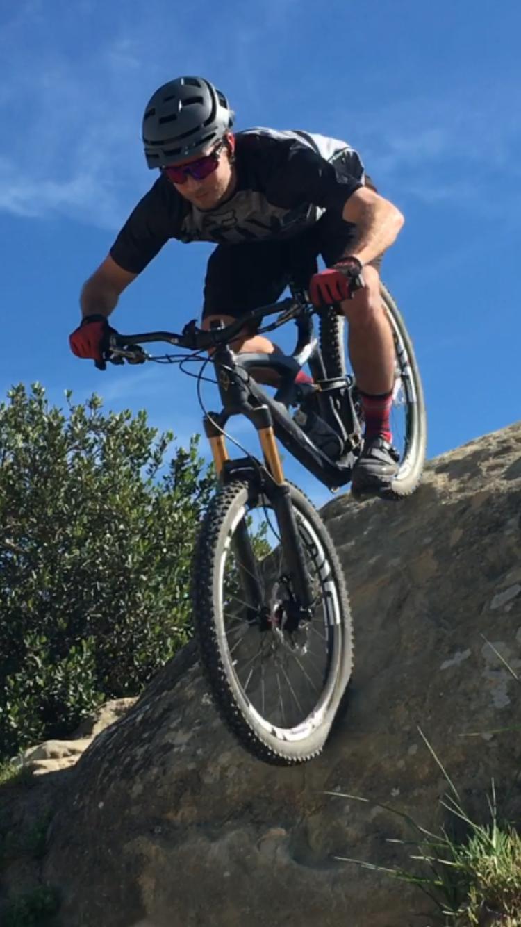 Ibis Mojo HD3: A mountain biker in a helmet and sunglasses descends a rocky outcrop, skillfully maneuvering their bike while surrounded by greenery and a clear blue sky.