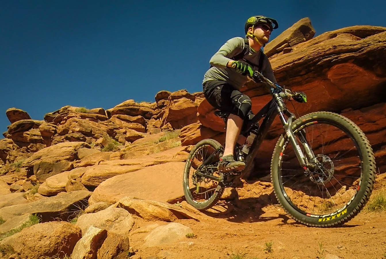 A mountain biker navigating a rocky trail, surrounded by red rocks under a clear blue sky. The biker is wearing a helmet and protective gear, showcasing an action-packed moment in outdoor sports. Captain Ahab mountain bike trail.