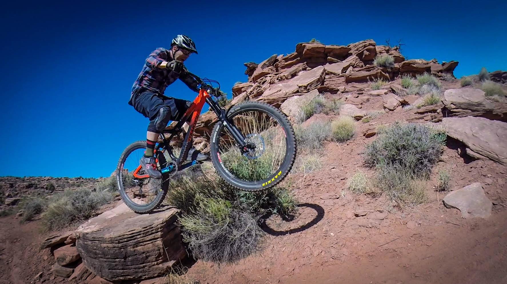 A mountain biker, wearing a helmet and protective gear, executes a jump over a rock formation in a desert landscape. The rider is dressed in a plaid shirt and shorts, with the bike's front wheel elevated off the ground, surrounded by sparse vegetation and reddish soil under a clear blue sky. Captain Ahab mountain bike trail.