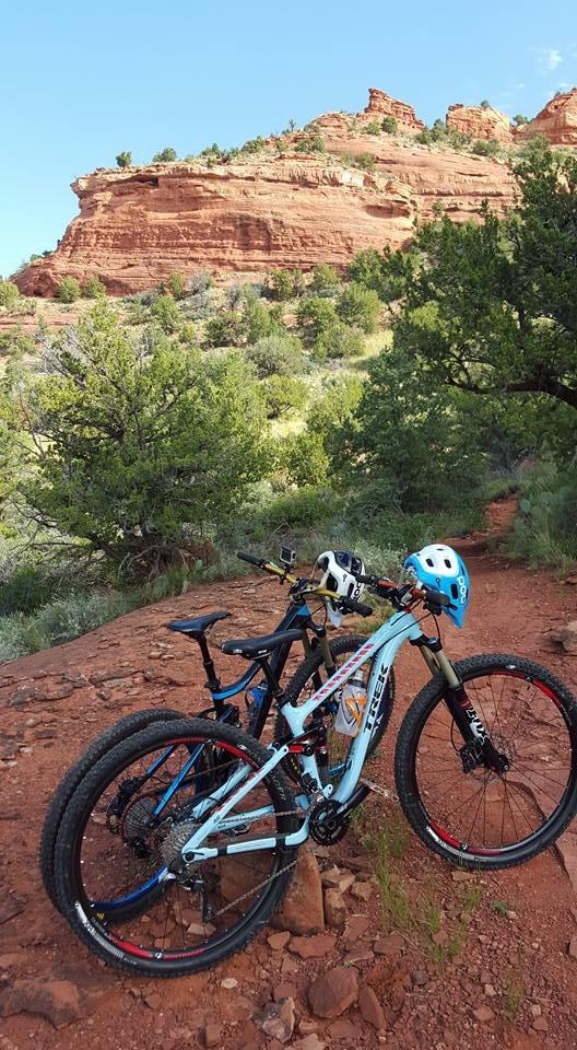 Trek Fuel EX 8: Two mountain bikes are leaning against a rock formation on a dirt trail surrounded by green shrubs and trees. In the background, there are red rock cliffs under a bright blue sky, indicating a scenic outdoor area suitable for biking.