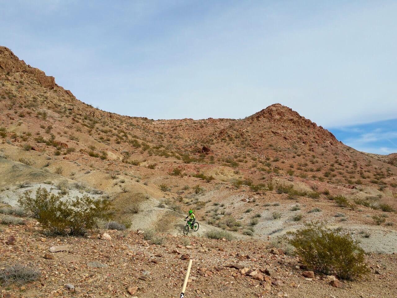 A mountain biker riding along rugged terrain with reddish-brown hills and sparse vegetation under a cloudy sky. Bootleg Canyon mountain bike trail.