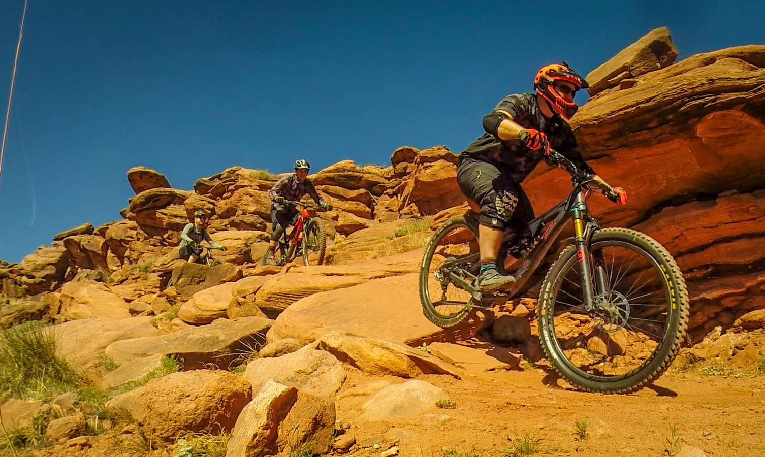 Three mountain bikers navigate rocky terrain under a clear blue sky. The scene showcases a mix of red rock formations and sandy paths, with the riders dressed in protective gear, demonstrating skill and excitement as they ride down the steep landscape. Captain Ahab mountain bike trail.