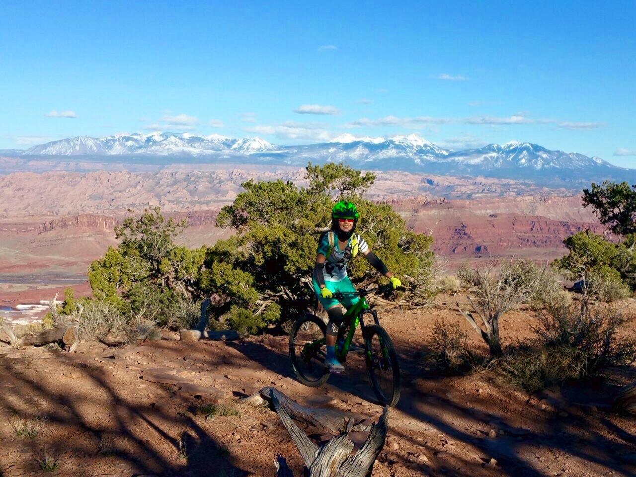 A mountain biker wearing a helmet and colorful gear stands beside their bike on a rocky trail, overlooking a stunning landscape adorned with mountains and blue sky. The scene features barren terrain with sparse vegetation and distant snow-capped peaks. Dead Horse Point State Park mountain bike trail.