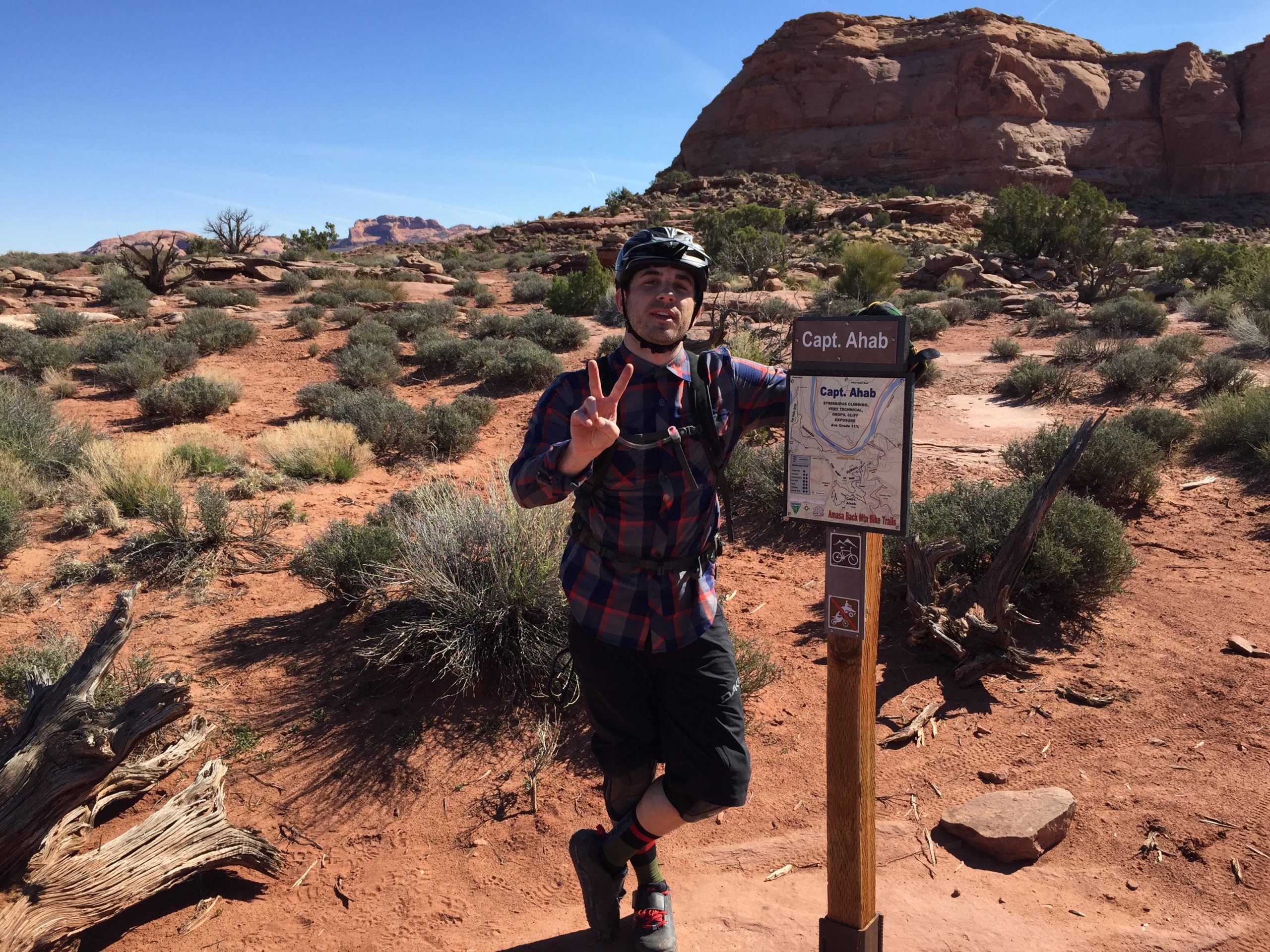 A person in a helmet and plaid shirt poses with a peace sign next to a trail sign labeled "Capt. Ahab" in a red rock landscape with sparse vegetation and blue sky in the background. Captain Ahab mountain bike trail.