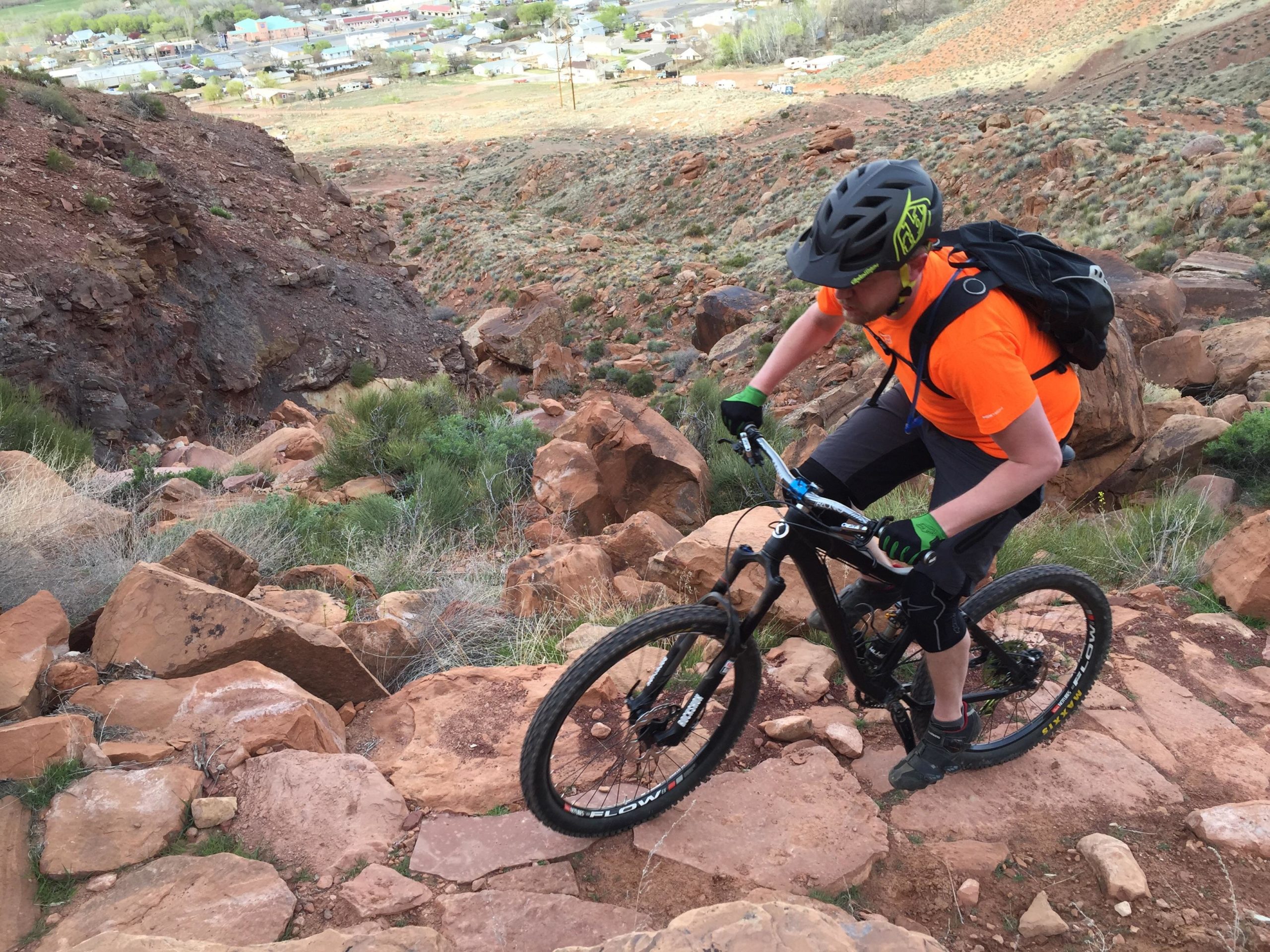 A mountain biker navigates a rocky terrain, dressed in an orange shirt and wearing a helmet, gloves, and a backpack. The landscape features reddish-brown rocks and sparse vegetation, with a residential area visible in the background. Pipe Dream mountain bike trail.
