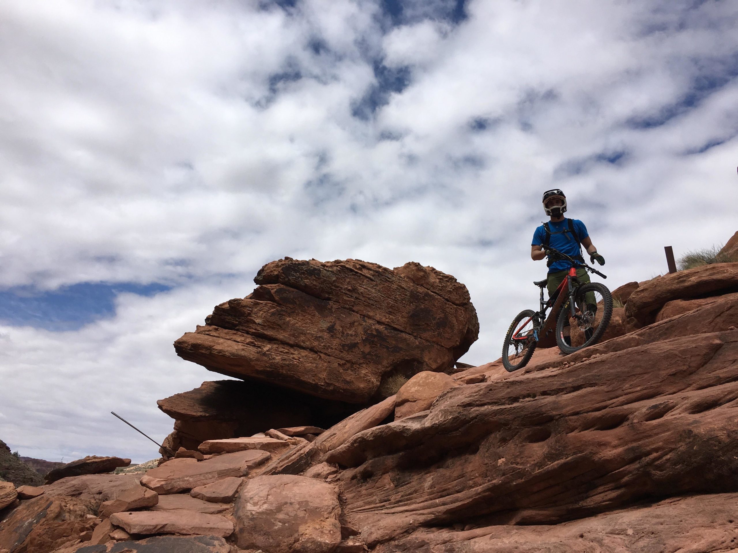 A mountain biker standing on rocky terrain with large boulders in the background, under a partly cloudy sky. The biker is wearing a helmet and riding gear, and is holding onto his bike while looking towards the camera. The landscape features red rock formations characteristic of desert environments. Porcupine Rim mountain bike trail.