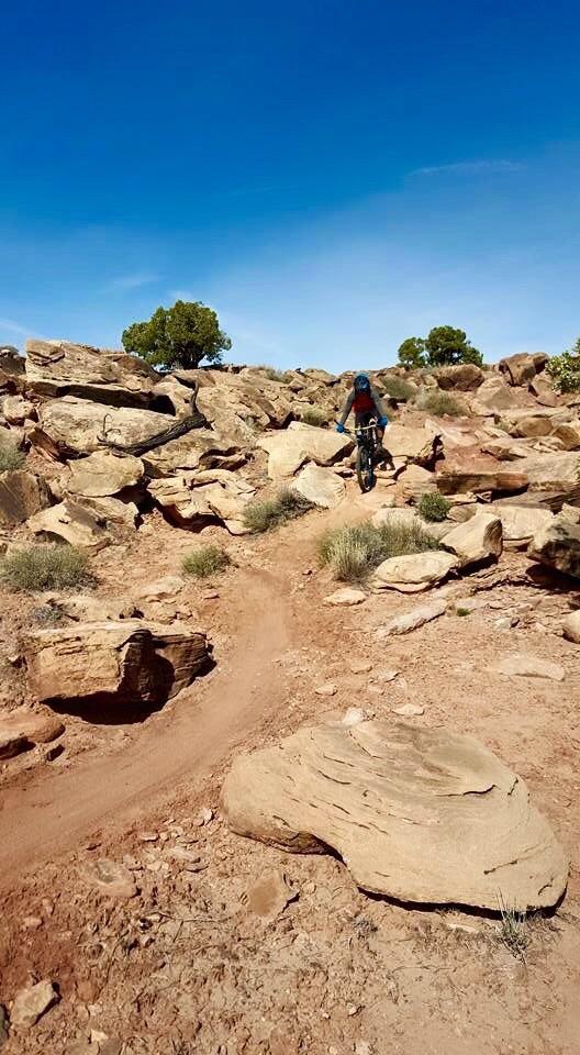 A mountain biker navigating a rocky trail in a desert landscape under a clear blue sky. The terrain features large boulders and sparse vegetation, with dirt paths winding through the rocks. Klondike Bluffs mountain bike trail.
