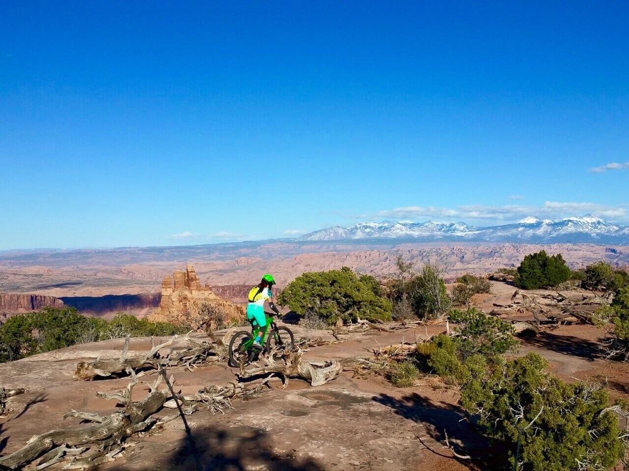 A mountain biker dressed in bright green rides along a rocky trail with expansive desert scenery in the background. The landscape features red rock formations, scattered trees, and distant snow-capped mountains under a clear blue sky. Dead Horse Point State Park mountain bike trail.