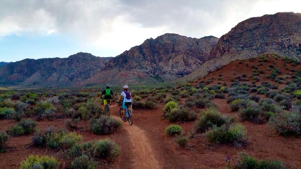 Two mountain bikers riding along a dirt path in a rocky landscape, surrounded by shrubs and vegetation, with mountains in the background under a partly cloudy sky. Blue Diamond mountain bike trail.