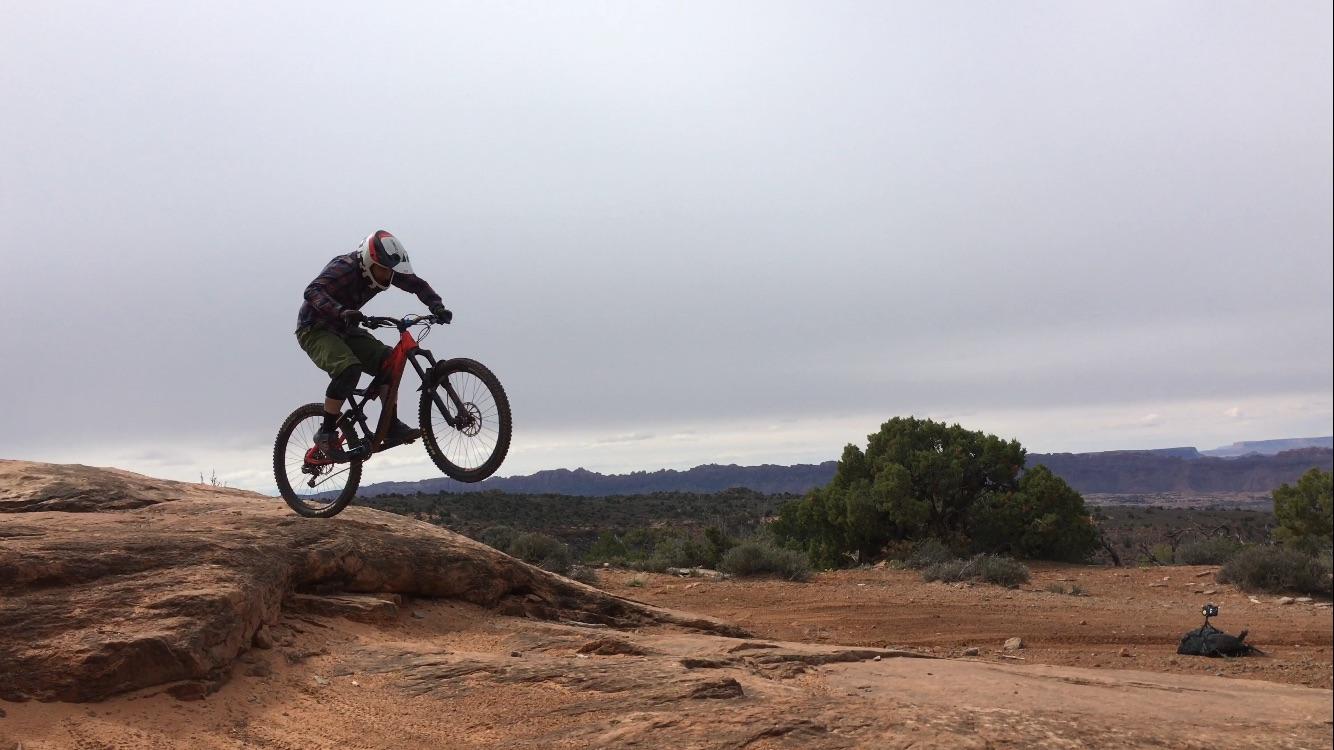 A mountain biker performing a jump over a rocky terrain with desert landscape and distant mountains in the background, under a cloudy sky. Porcupine Rim mountain bike trail.