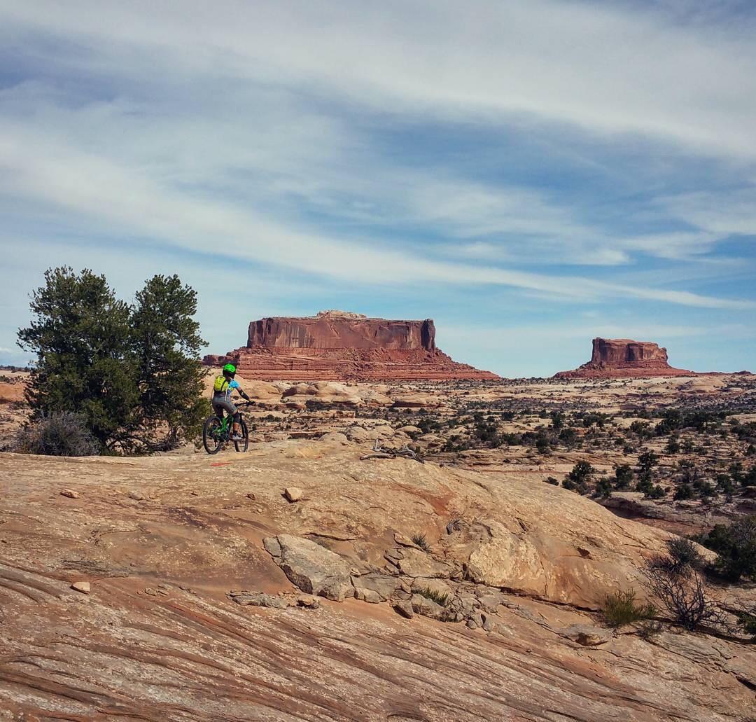 A cyclist in a green helmet is riding on rocky terrain, with large mesas rising in the background under a clear blue sky. Scrub vegetation is visible amidst the rugged landscape, showcasing the natural beauty of the area. Navajo Rocks mountain bike trail.