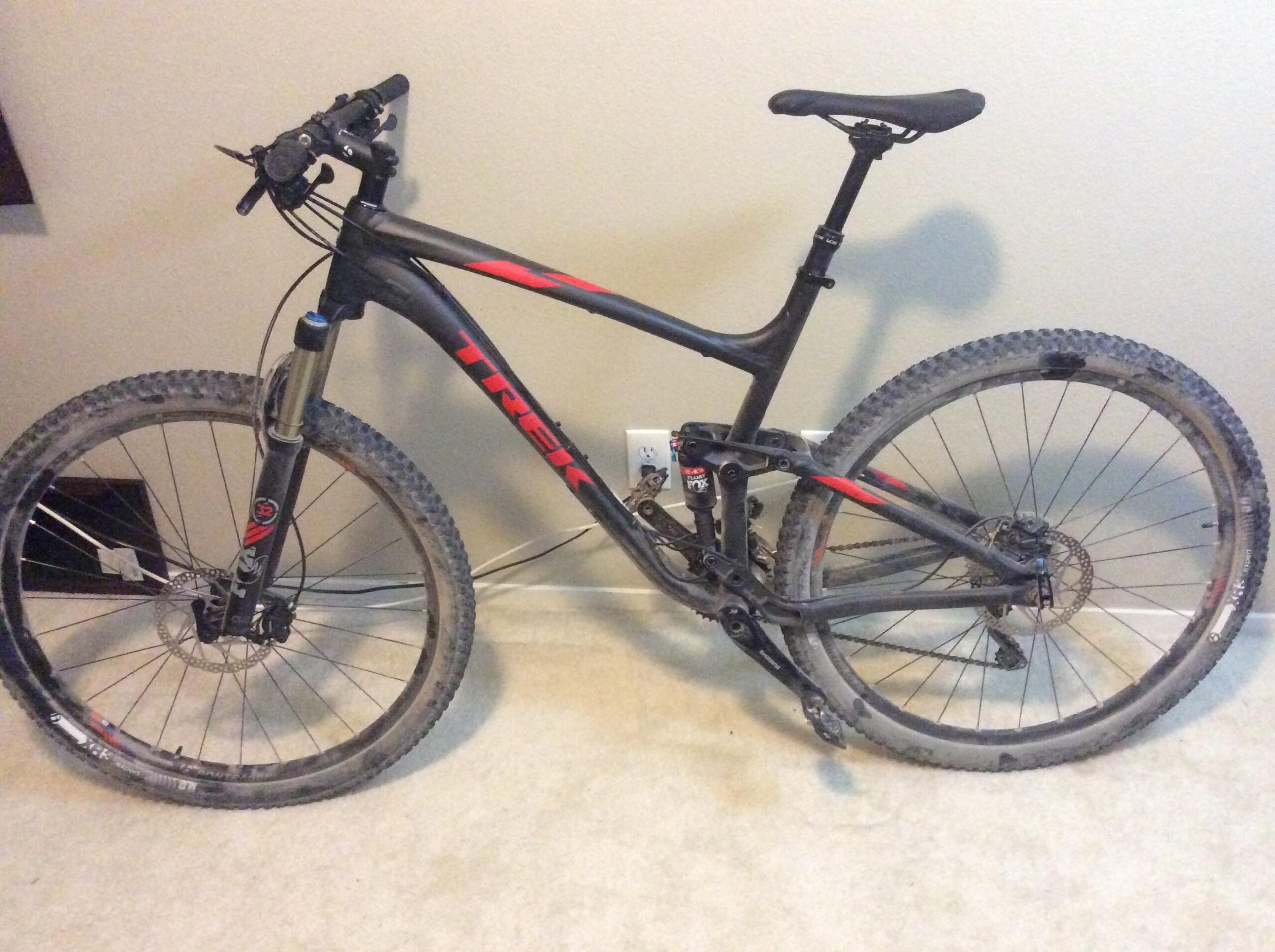 Trek Fuel EX 8: A black and red Trek mountain bike leaning against a wall, with visible dirt on the tires and frame, set on a carpeted floor.