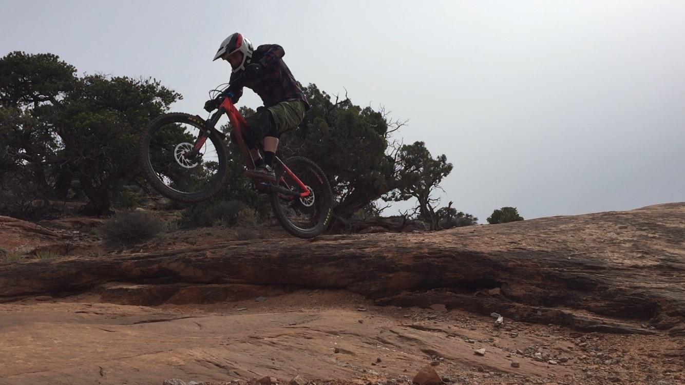 A mountain biker performing a jump on a rugged trail, with a rocky terrain and sparse vegetation in the background. The rider is wearing a helmet and protective gear, and is airborne with their bike angled above a large rock. Porcupine Rim mountain bike trail.