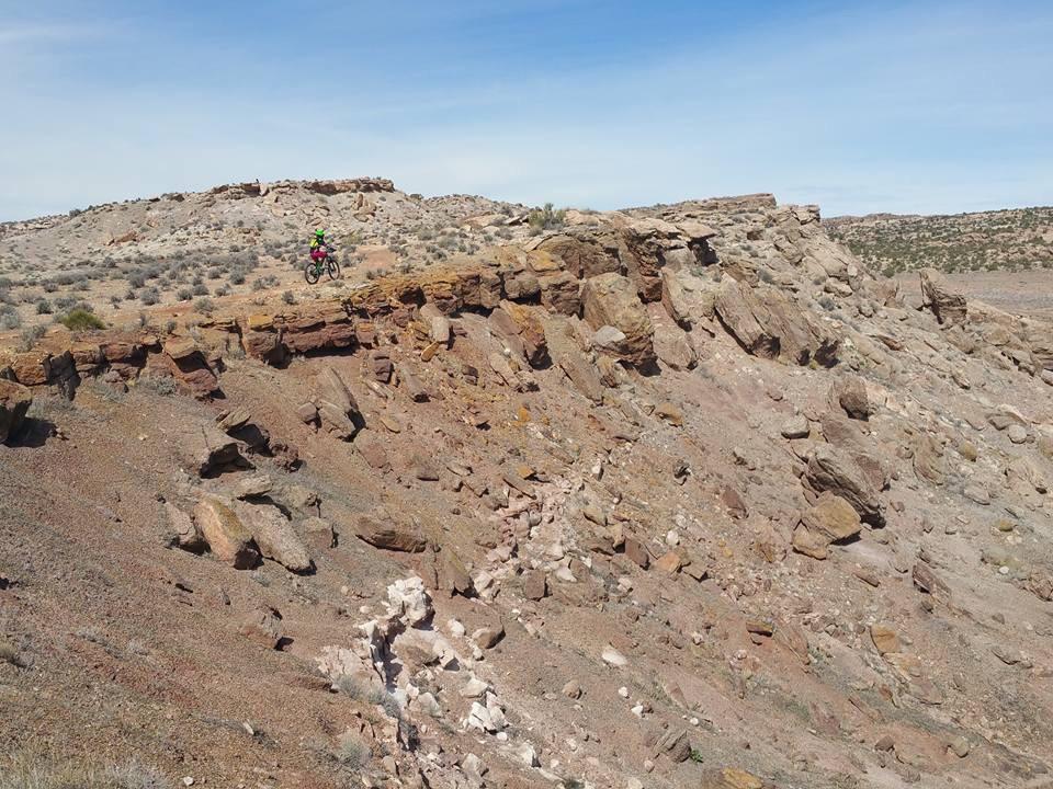 A person riding a mountain bike along the edge of a rocky cliff in a desert landscape, surrounded by sparse vegetation and dramatic terrain under a clear blue sky. Klondike Bluffs mountain bike trail.