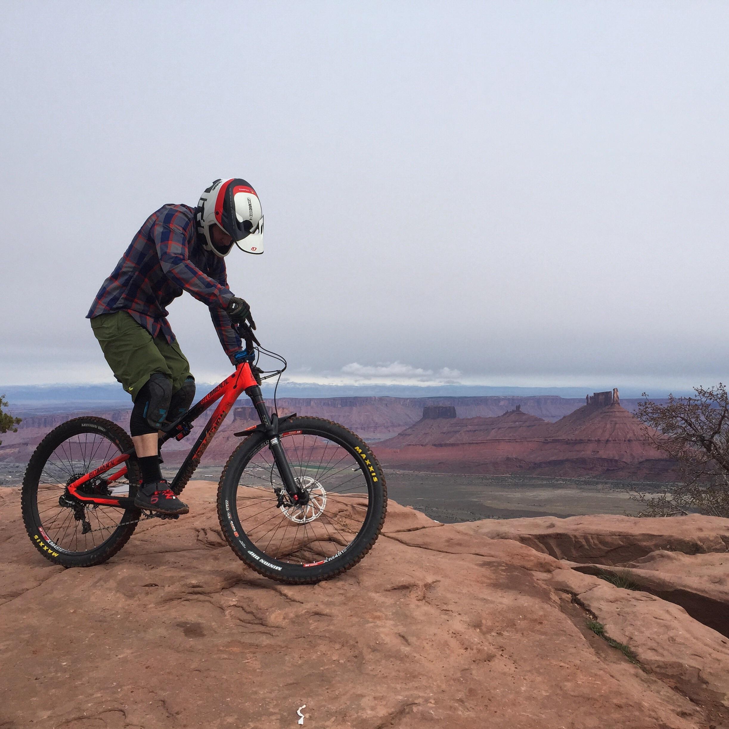 A mountain biker on a rocky ledge, wearing a helmet and protective gear, balances on a red bicycle. The backdrop features a vast landscape of red rock formations and distant mountains under a cloudy sky. Porcupine Rim mountain bike trail.