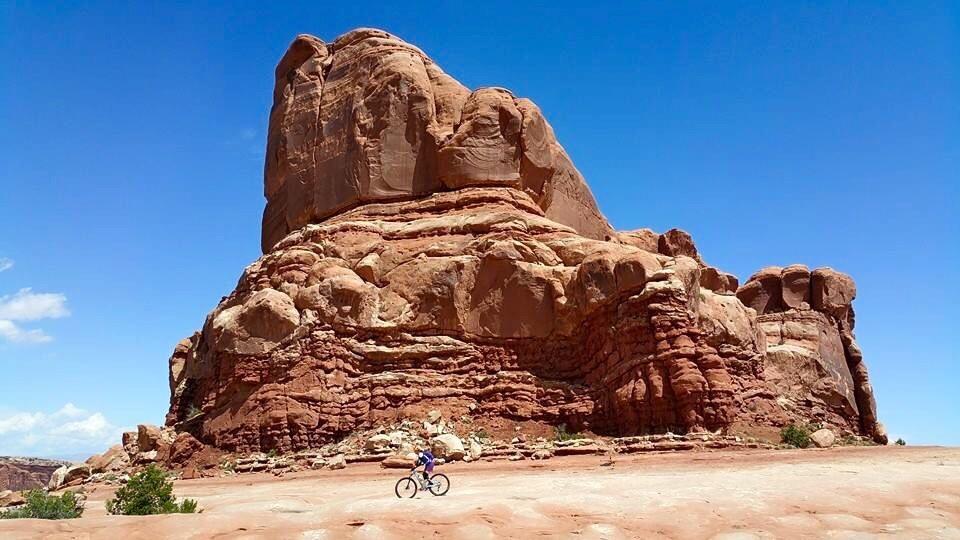 A cyclist rides along a sandy path in front of a large, reddish rock formation under a clear blue sky. Scarce vegetation is visible near the base of the rock, highlighting the natural landscape. Monitor & Merrimac Singletrack mountain bike trail.