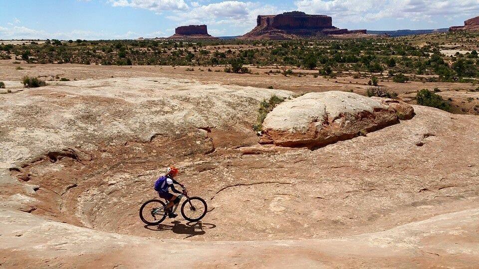 A person riding a mountain bike on a rocky terrain with desert landscape in the background, featuring mesas and a clear blue sky with scattered clouds. Monitor & Merrimac Singletrack mountain bike trail.