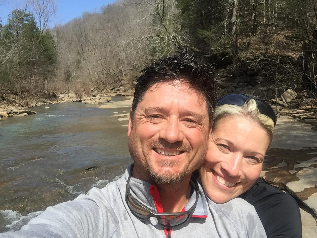 A smiling couple poses for a selfie in a natural outdoor setting by a stream, with trees and rocky terrain in the background. The sun is shining, and both individuals appear happy and relaxed. Fossil Flats mountain bike trail.
