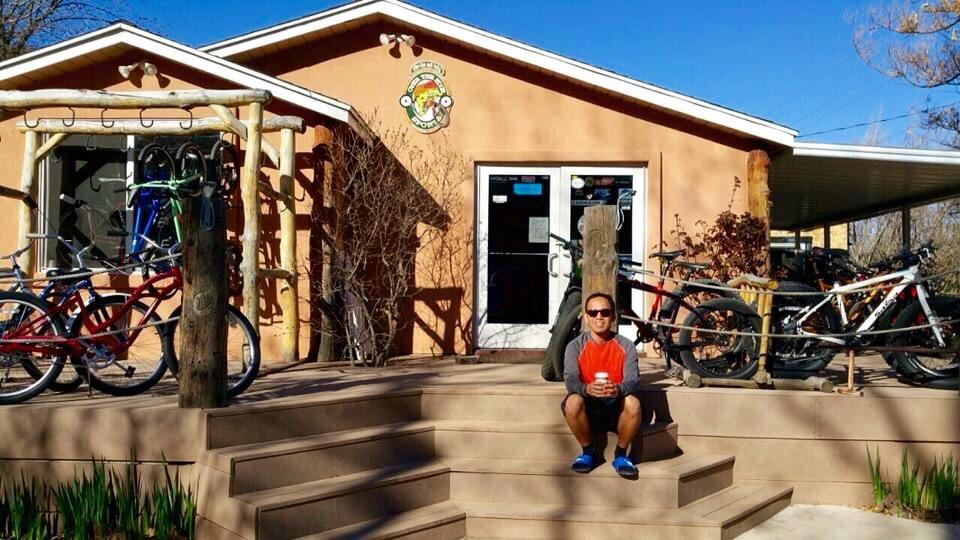 A person sitting on the steps of a bike rental shop, surrounded by parked bicycles. The building is light-colored with large windows and a logo displayed above the entrance, under a clear blue sky. Trees and greenery are visible in the foreground, creating a relaxed outdoor atmosphere.