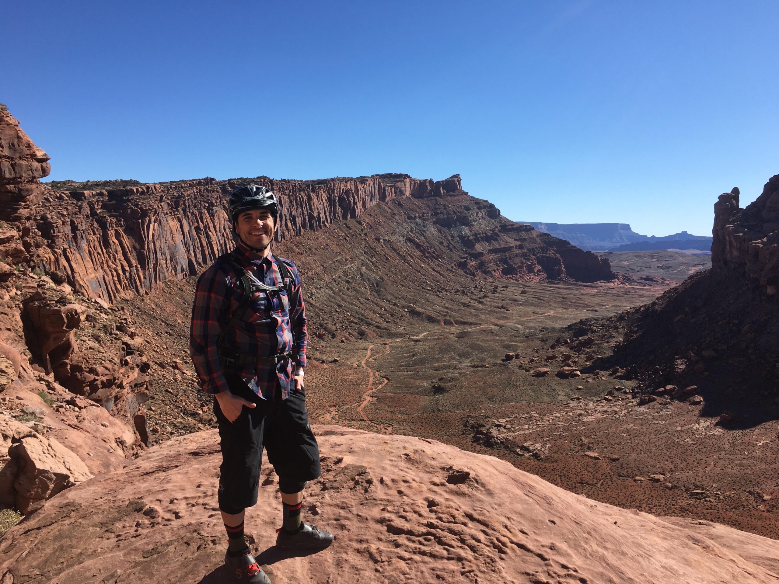 A person wearing a helmet and outdoor attire stands on a rocky outcrop in a desert landscape, smiling at the camera. In the background, towering red rock formations and a vast, arid valley stretch under a clear blue sky. Amasa Back Trail mountain bike trail.