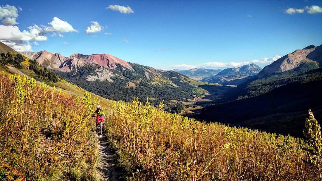 A cyclist rides along a winding dirt trail surrounded by vibrant autumn foliage, with expansive mountain ranges in the background. The sky is clear with a few clouds, creating a picturesque landscape. Trail 401 mountain bike trail.